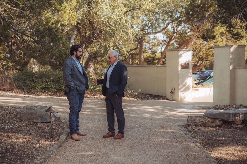 Two people stand on a concrete path surrounded by autumn trees and fallen leaves in an outdoor setting.