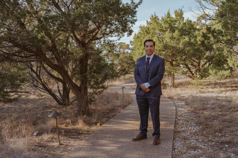A person in a navy blue suit stands on a dirt path surrounded by desert brush and pine trees.
