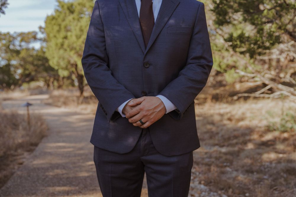 A person in a dark navy suit stands outdoors against a natural wooded background on a sunny day.