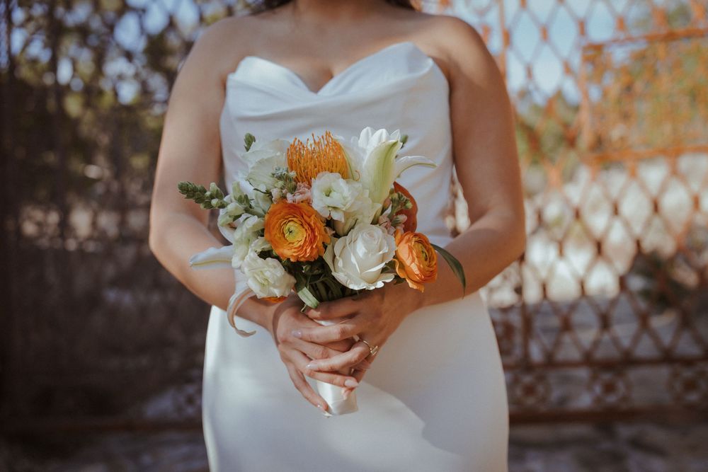 A bridal bouquet featuring orange and white flowers is held against a white dress with a rustic backdrop.