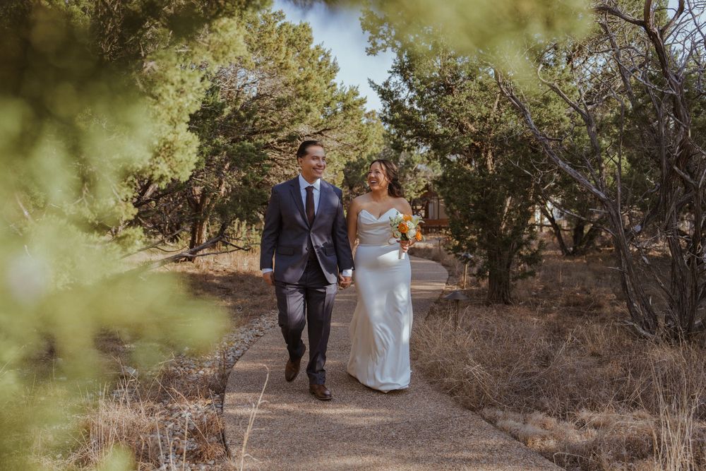 A couple walks hand in hand along a rustic dirt path surrounded by desert brush and trees on their wedding day.