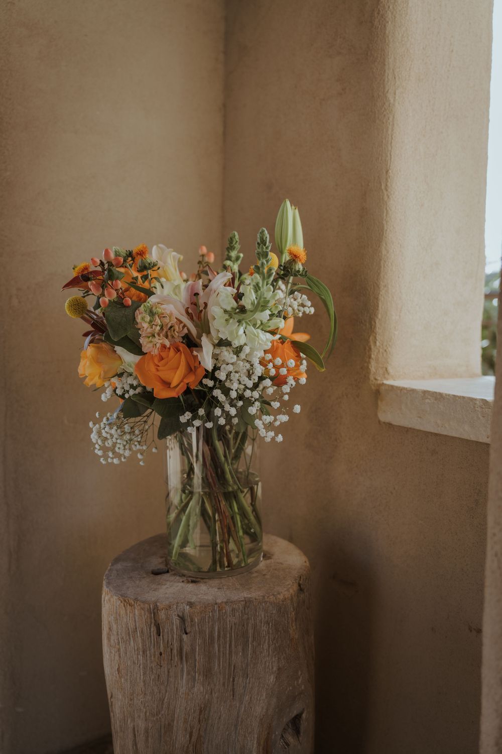 A rustic floral arrangement with orange roses and baby's breath in a glass vase sits atop a wooden pedestal.