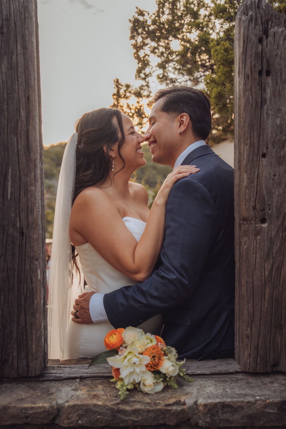 Newlywed couple shares intimate moments through rustic wooden posts with autumn bouquet in foreground at sunset.