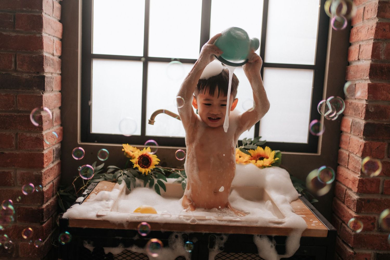 A child plays in a bubble-filled bath near a window with sunflowers, pouring water from a green cup.