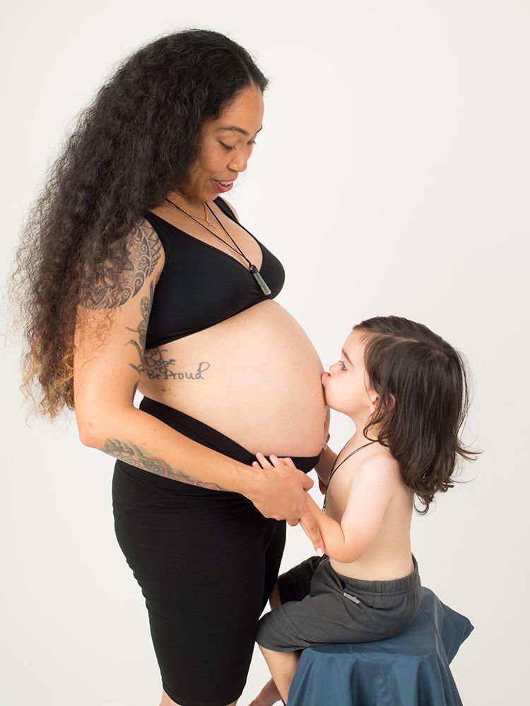 A pregnant woman in a black outfit stands sideways, smiling as a young child gently kisses her belly. Maternity photographer High Wycombe