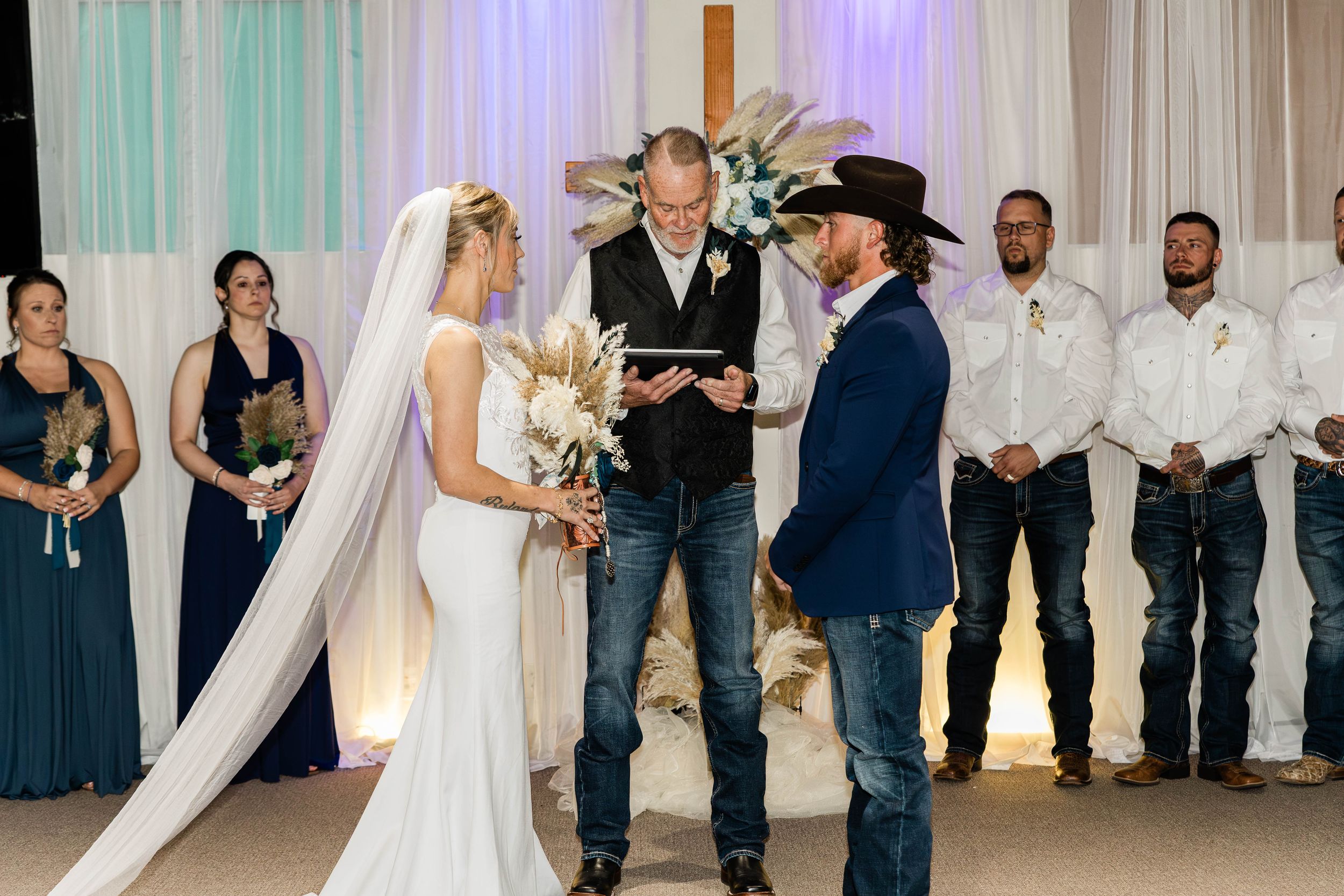 Country wedding ceremony with couple at altar surrounded by wedding party in blue attire.