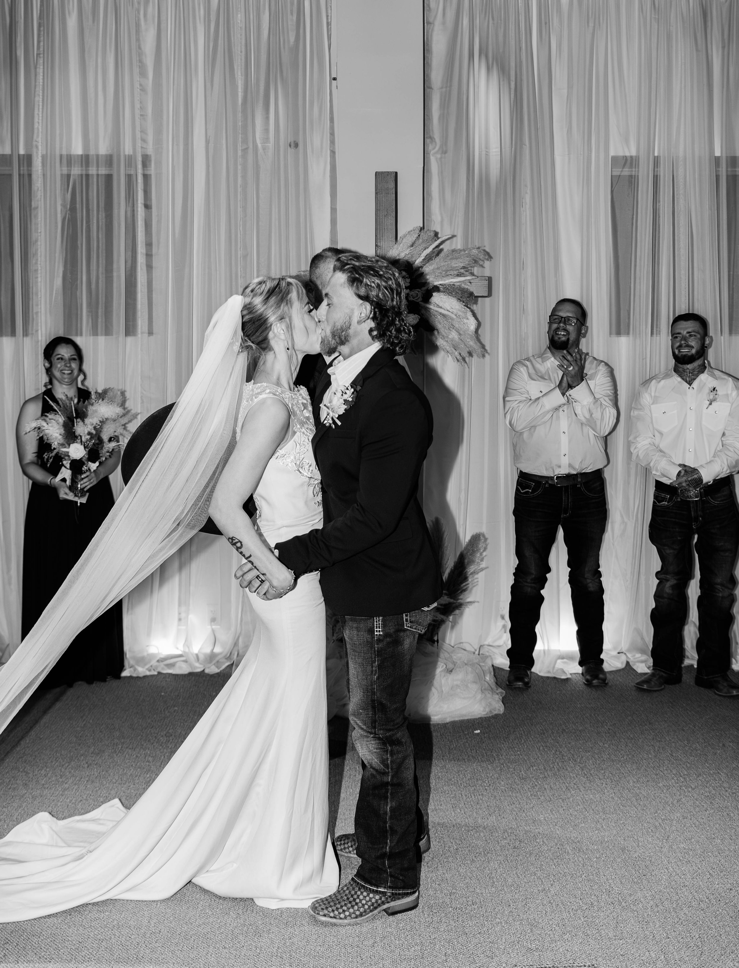 A romantic first kiss between newlyweds at their wedding ceremony with guests looking on.