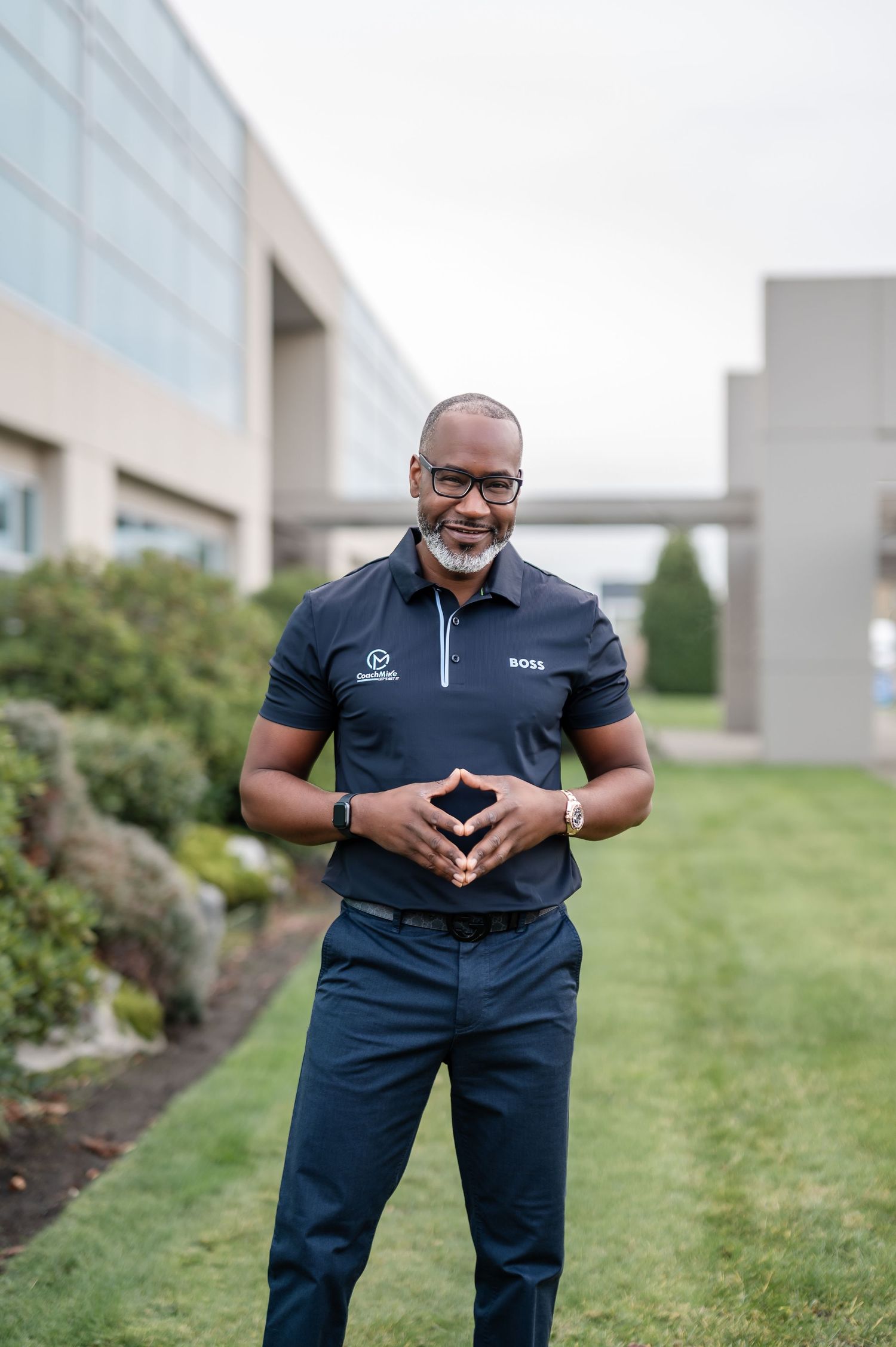 Professional wearing a navy blue polo shirt and slacks stands outdoors on grass near a modern building exterior.