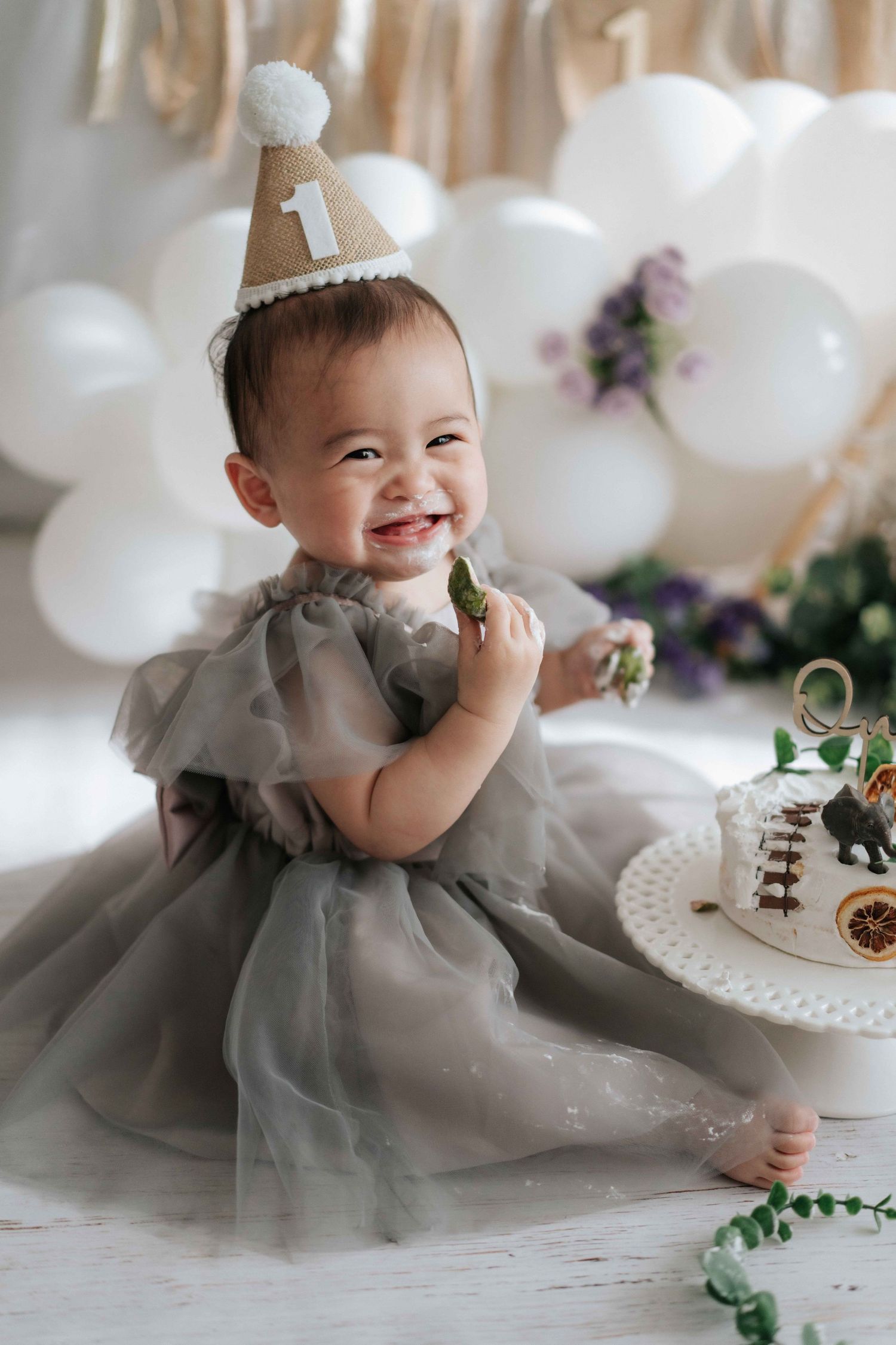 Baby wearing gray tulle dress smiles while sitting on floor with white balloons and floral decorations at party.
