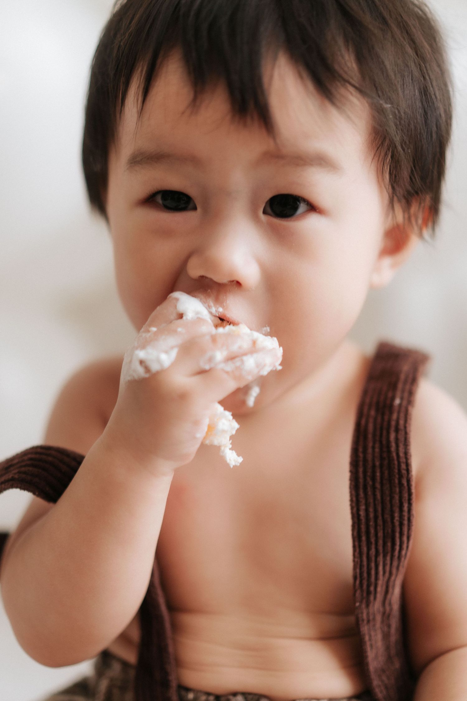 A small child in brown overalls enjoys eating a messy dessert with white cream or frosting on their face.