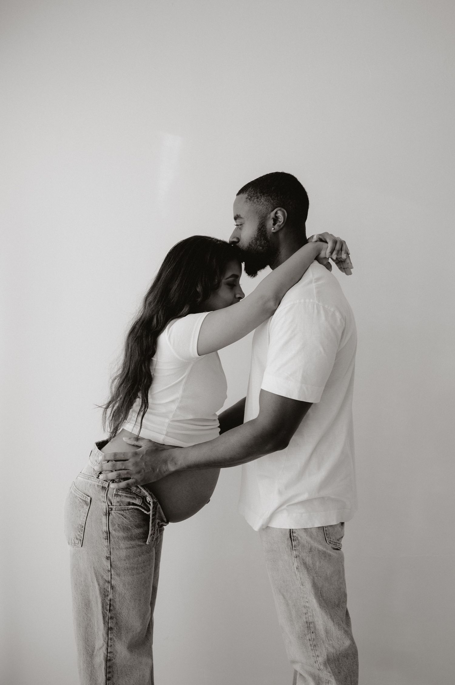 A couple in white shirts and jeans embrace in an artistic black and white photo series against a plain backdrop.