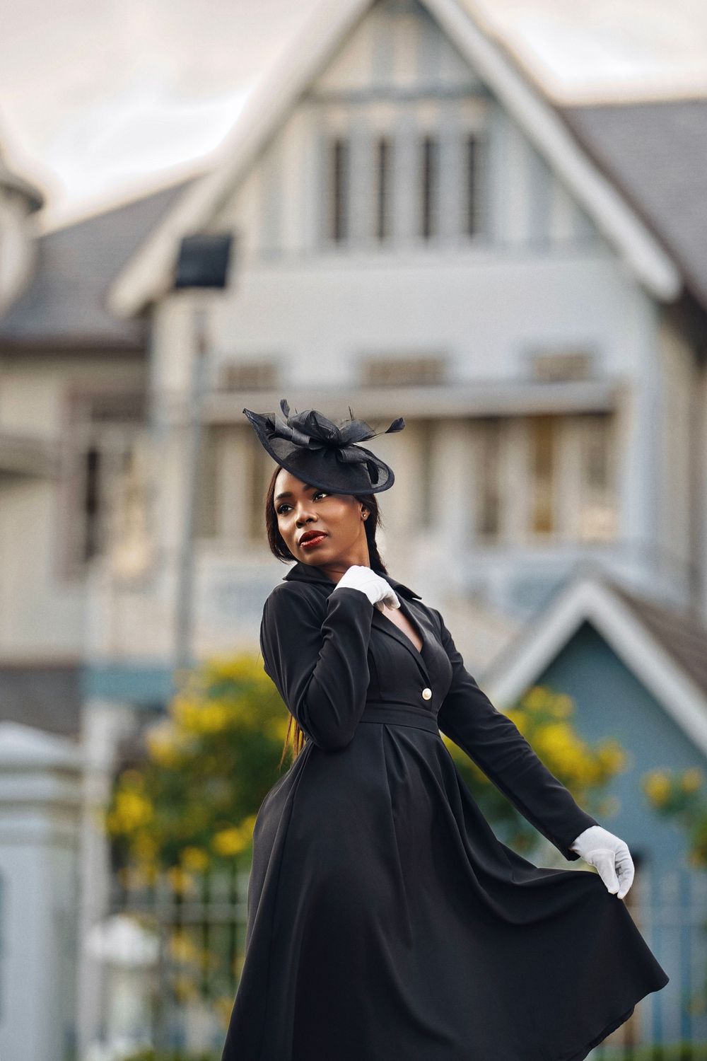 A figure in a flowing black dress and fascinator strikes dynamic poses against a backdrop of suburban houses.
