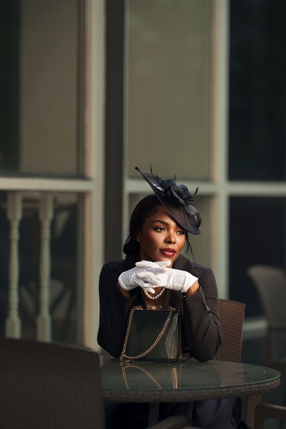 A fashionable person in a black blazer and white gloves poses elegantly at an outdoor cafe table.
