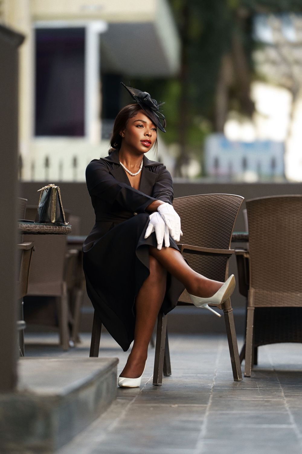 An elegant figure in a black ensemble and white gloves poses gracefully on outdoor furniture.