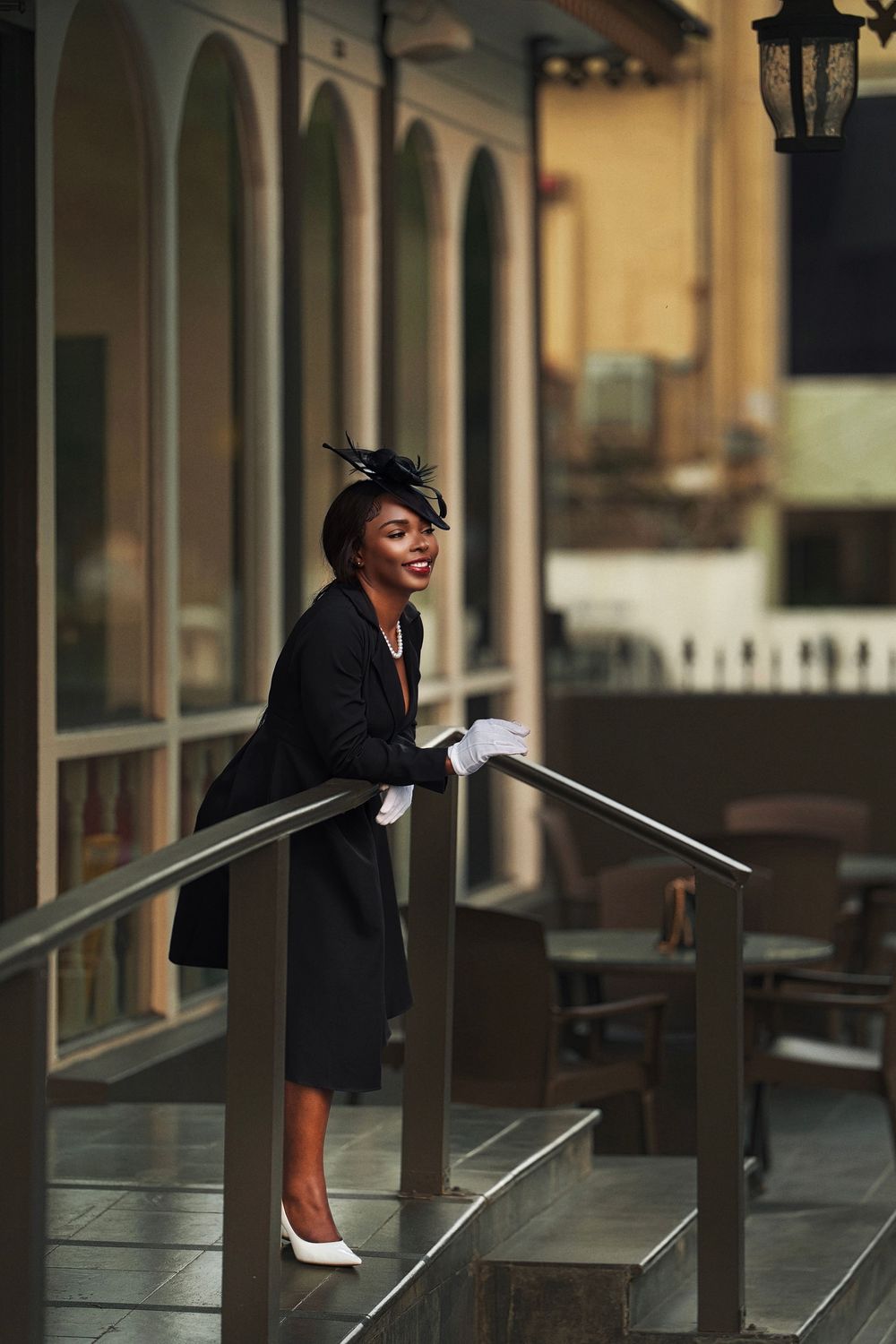 A person in a black blazer leans on a railing in an outdoor corridor with warm lighting and architectural columns.