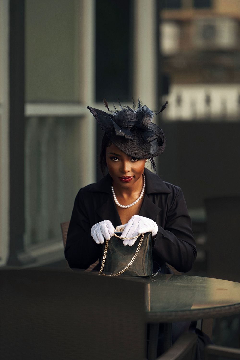 A stylish figure in black attire and pearls poses elegantly against a railing in a covered walkway.