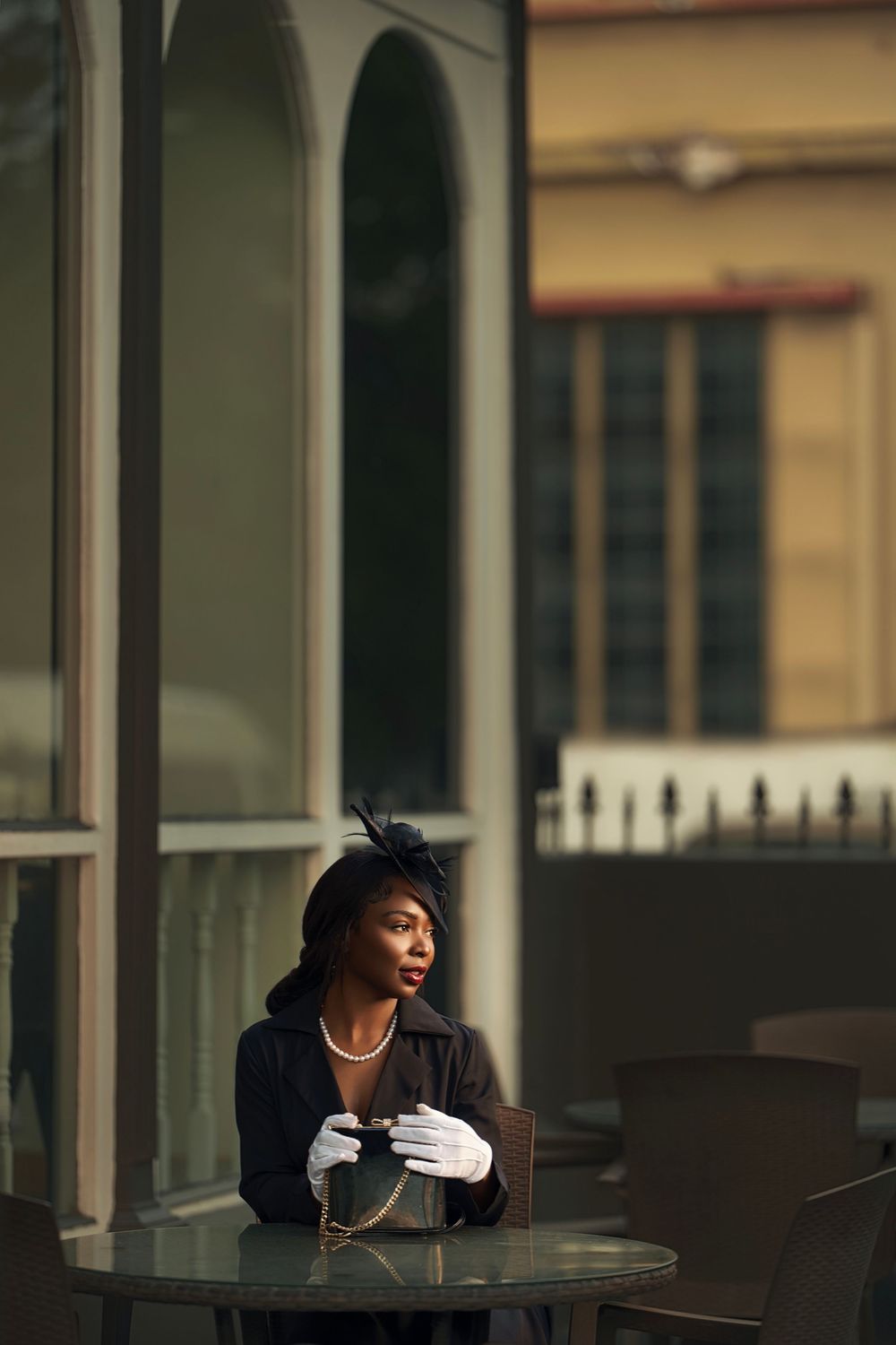 A figure in dark attire sits contemplatively at a table with white gloves and pearl accessories.