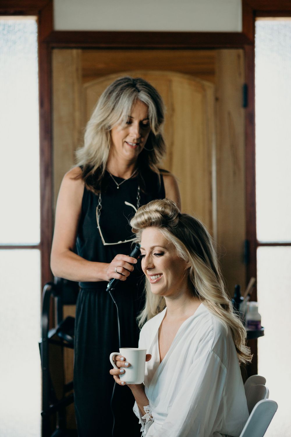 Hairstylist working on a bride's hair during wedding preparations in a warmly lit room.