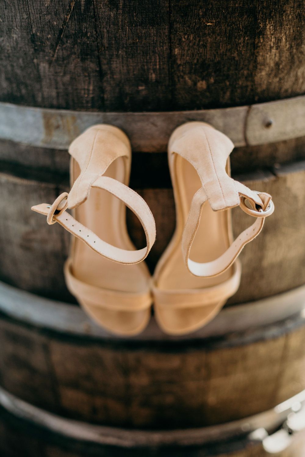 Elegant nude-colored sandals with ankle straps resting on a rustic wooden wine barrel.