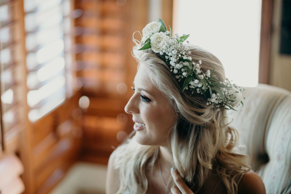 A bride and bridesmaids wearing floral crowns get ready in a sunlit room with wooden shutters.
