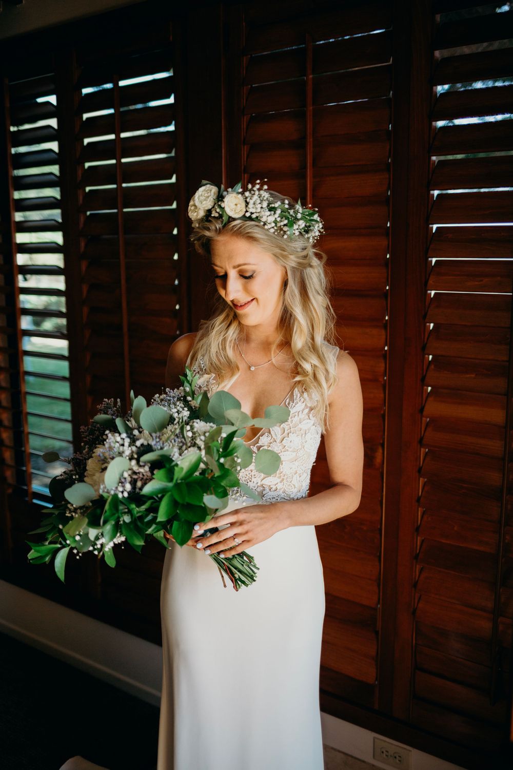 A bride in a white lace gown holds a bouquet of white and green flowers against wooden shutters.