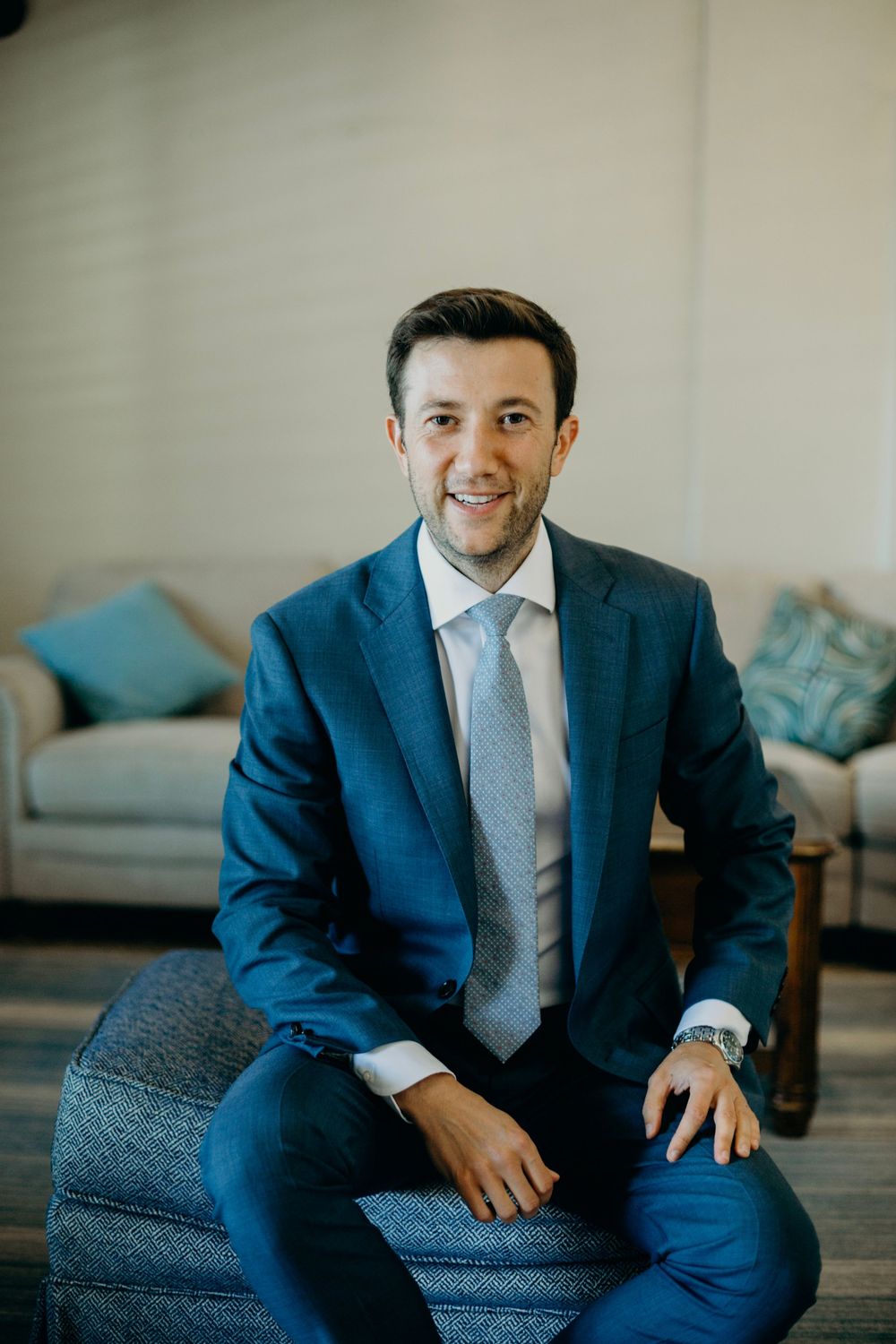 A groom in a navy blue suit and gray tie sits on a blue chair in a living room setting.