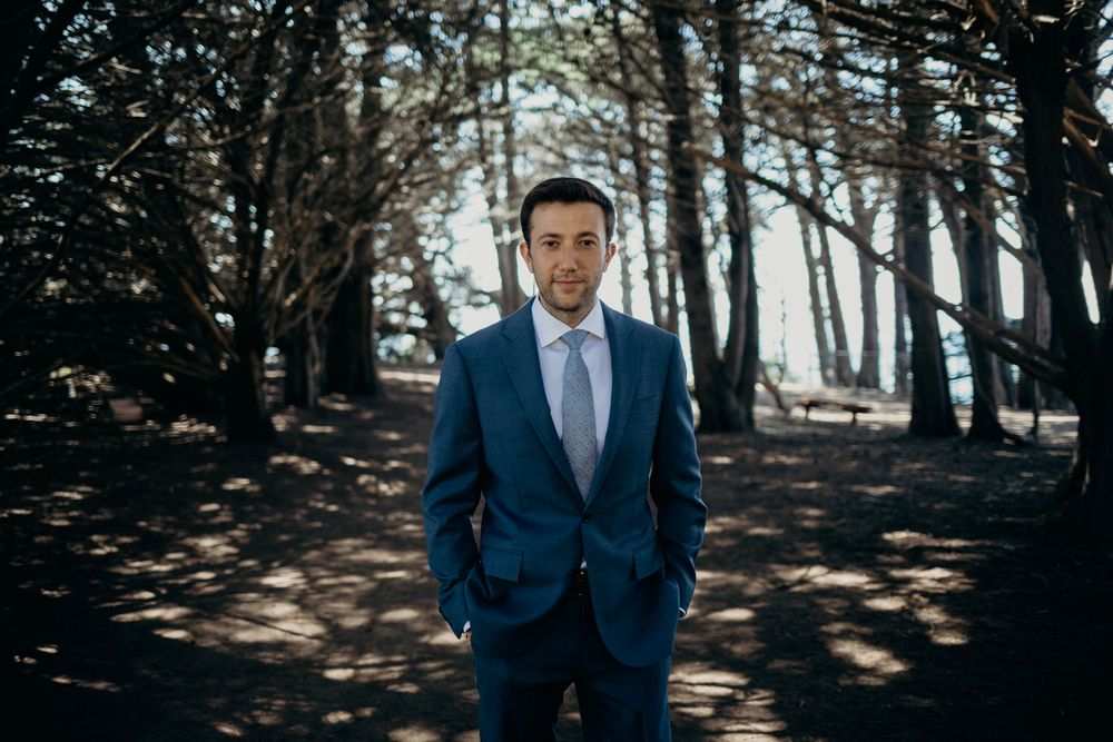 A person in a navy suit and blue tie stands in a sunlit forest with dappled light filtering through the trees.
