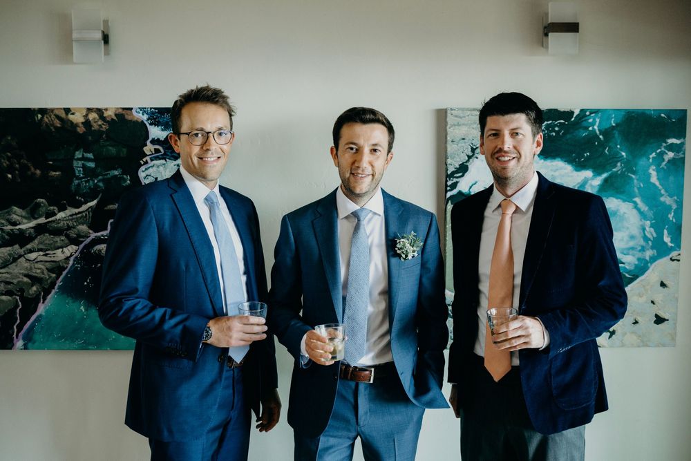 Three groomsmen in navy suits share a drink while getting ready for a wedding celebration.