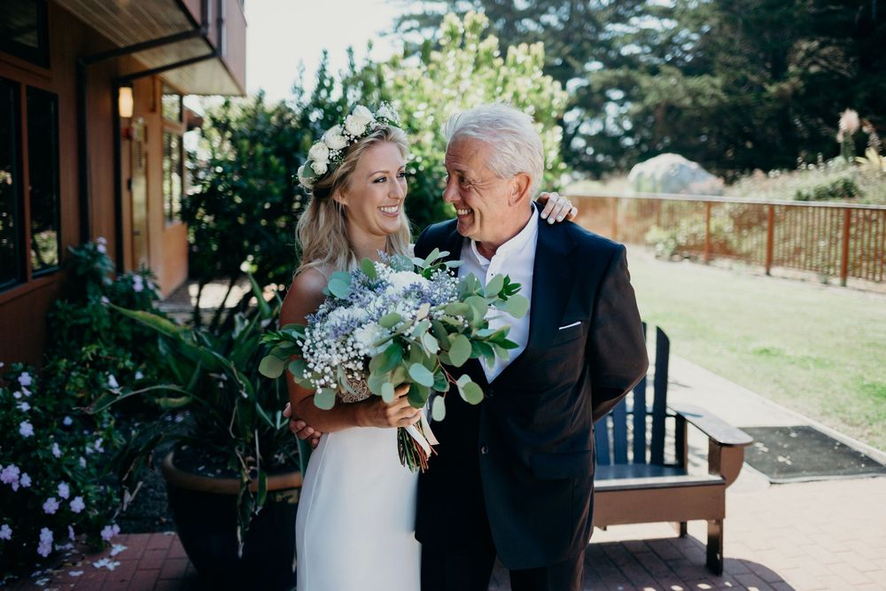 A bride in a white dress holds a bouquet of blue and green flowers while sharing a tender moment outdoors.