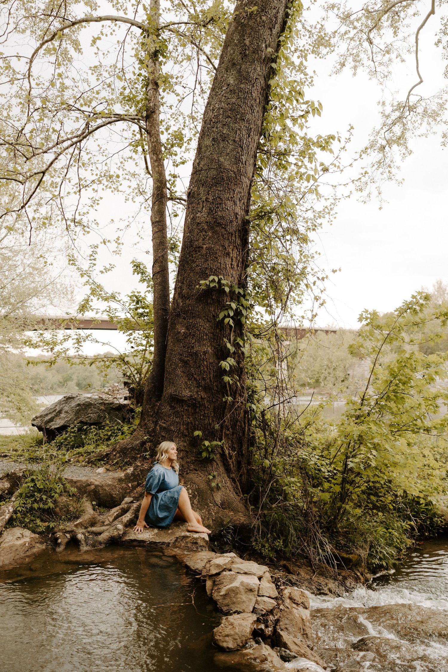 Woman in a teal dress sits peacefully on rocks beside a flowing woodland stream.
