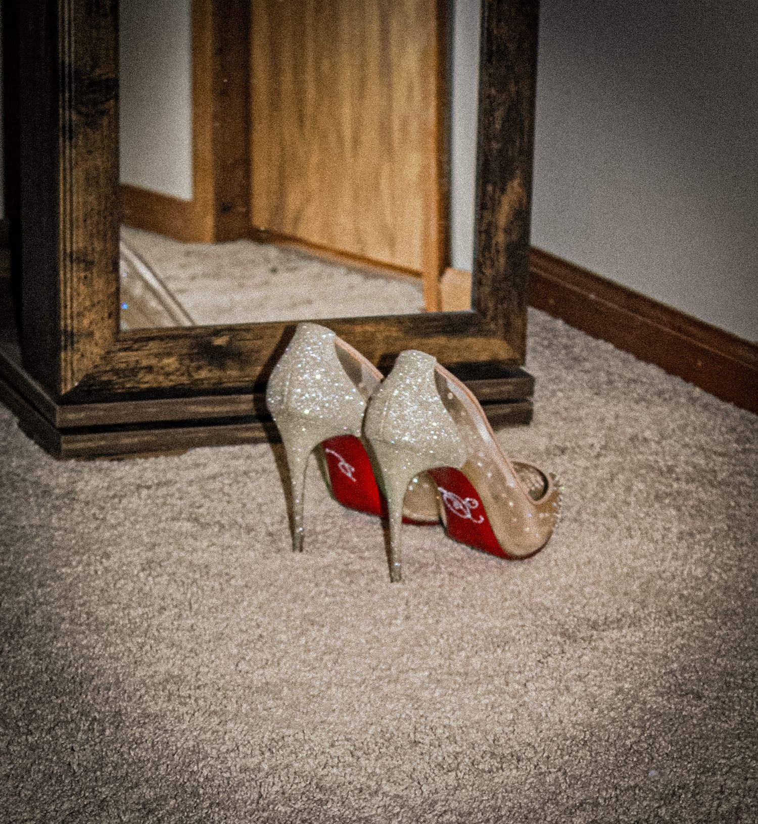 A pair of glittery silver high heel shoes with red soles sits on carpet next to a wooden mirror frame.