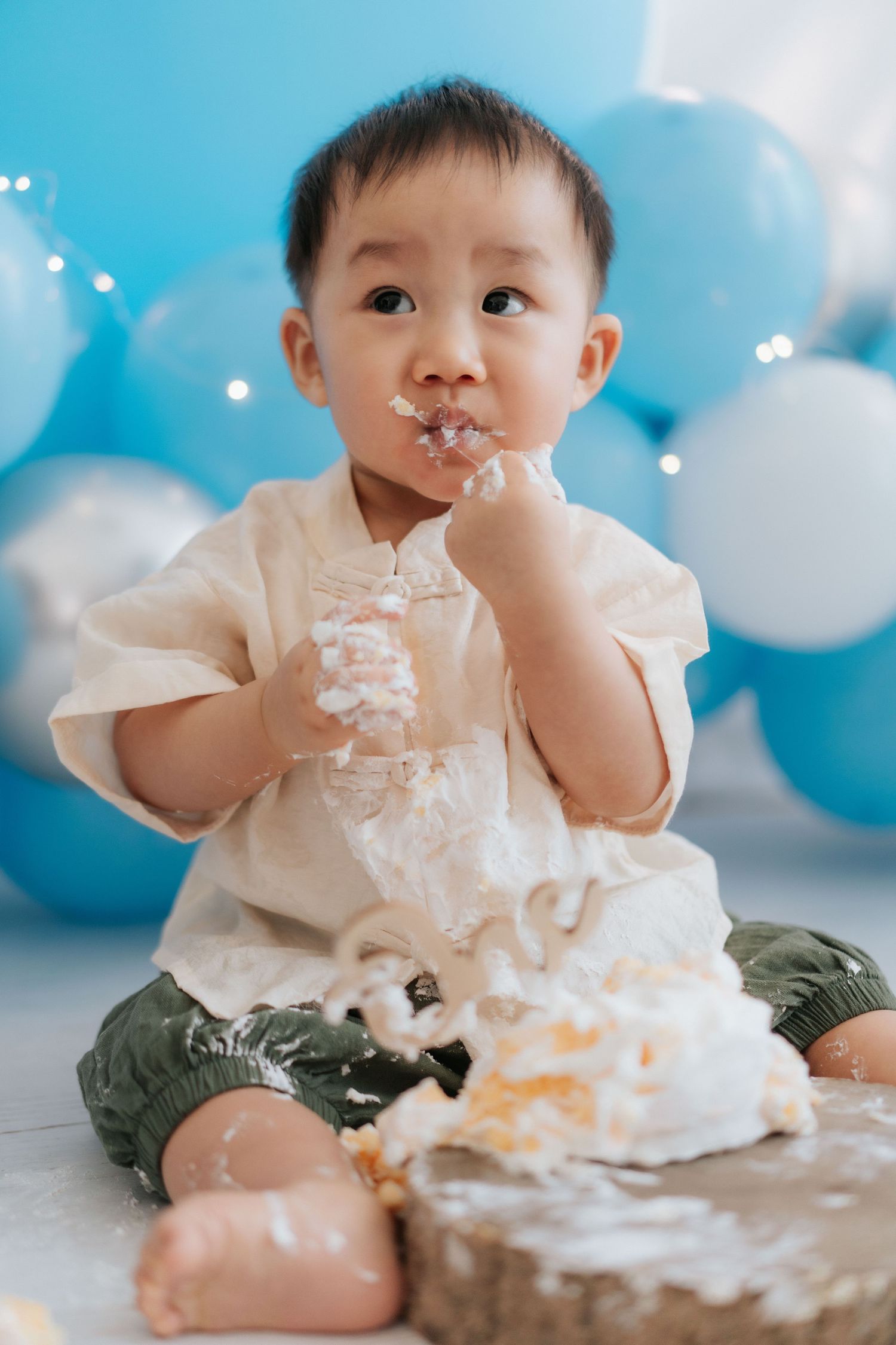A cake smash photo session featuring a baby in a white dress surrounded by balloons and decorative elements.