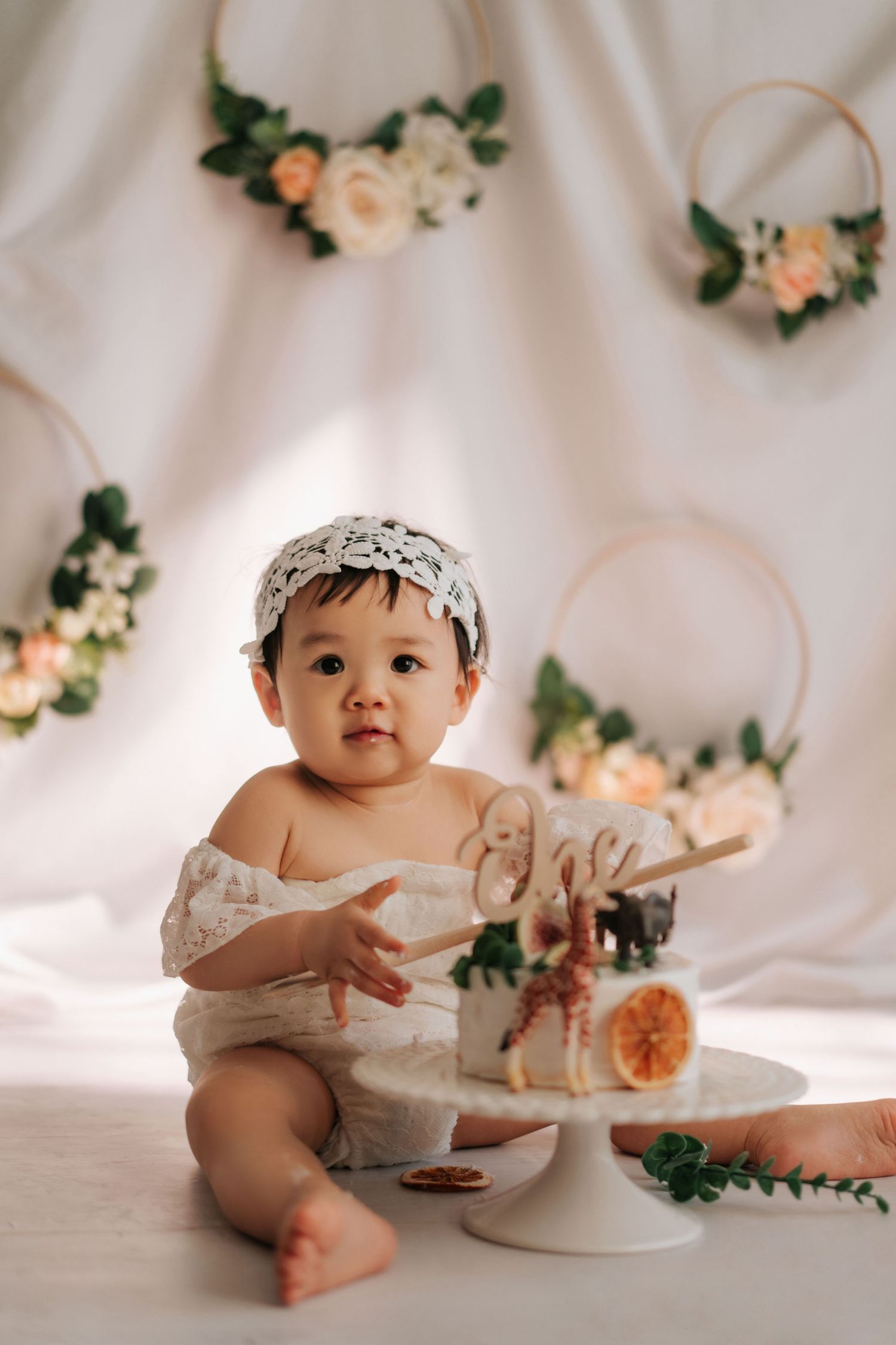 Baby in white outfit having a cake smash photo session with peach roses and greenery decorations against white backdrop.