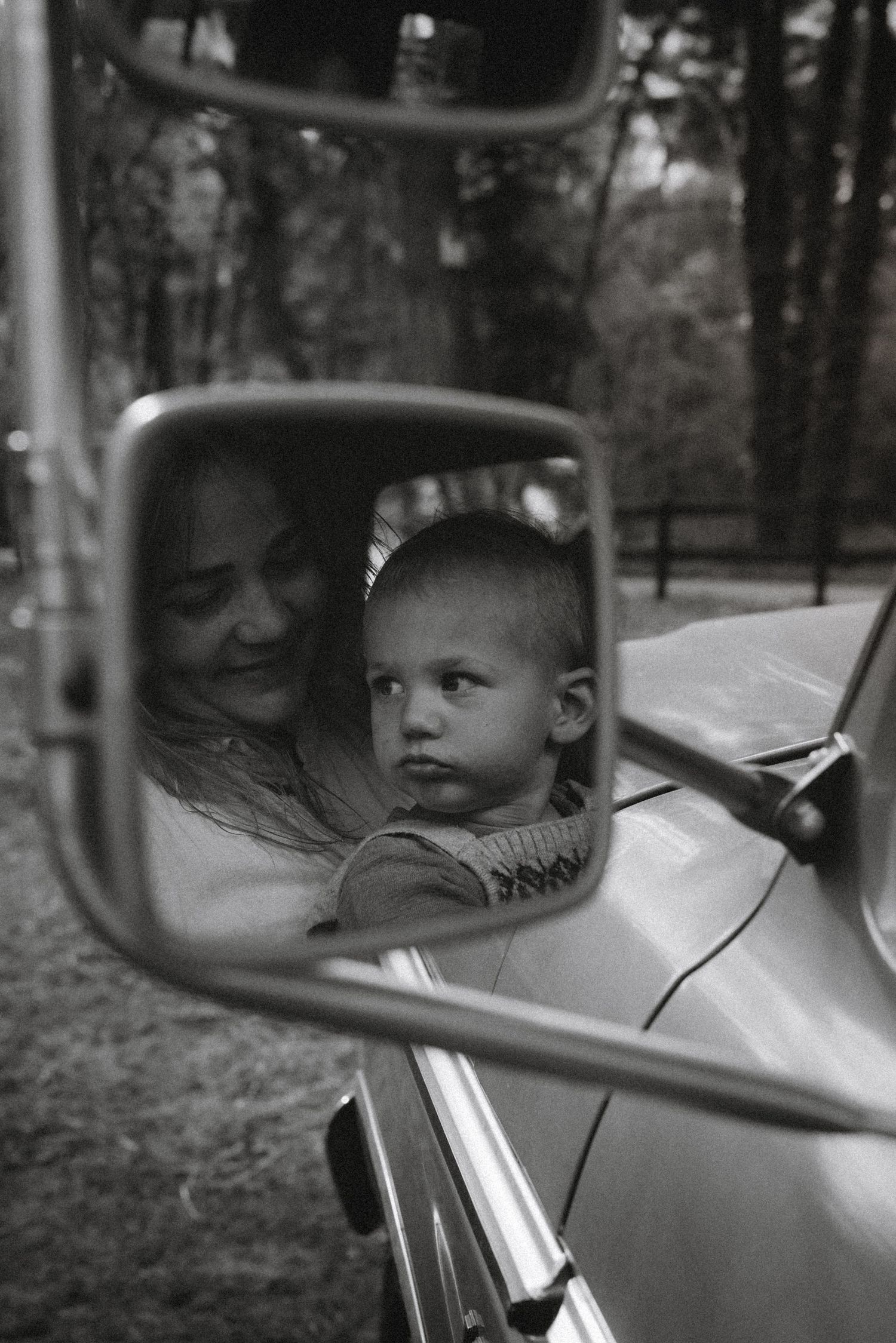 Black and white photo of mother and baby reflected in a vintage car side mirror, surrounded by trees. Javea photographer