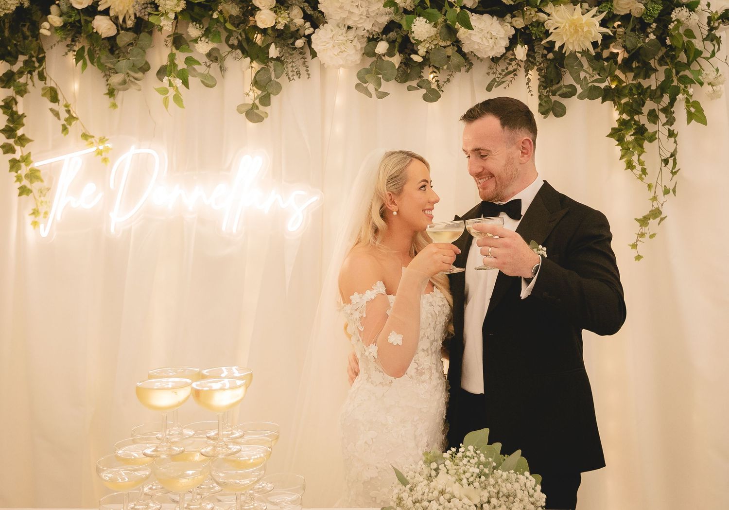 A romantic wedding celebration scene with dancing and neon lights under elegant greenery decor and warm lighting.