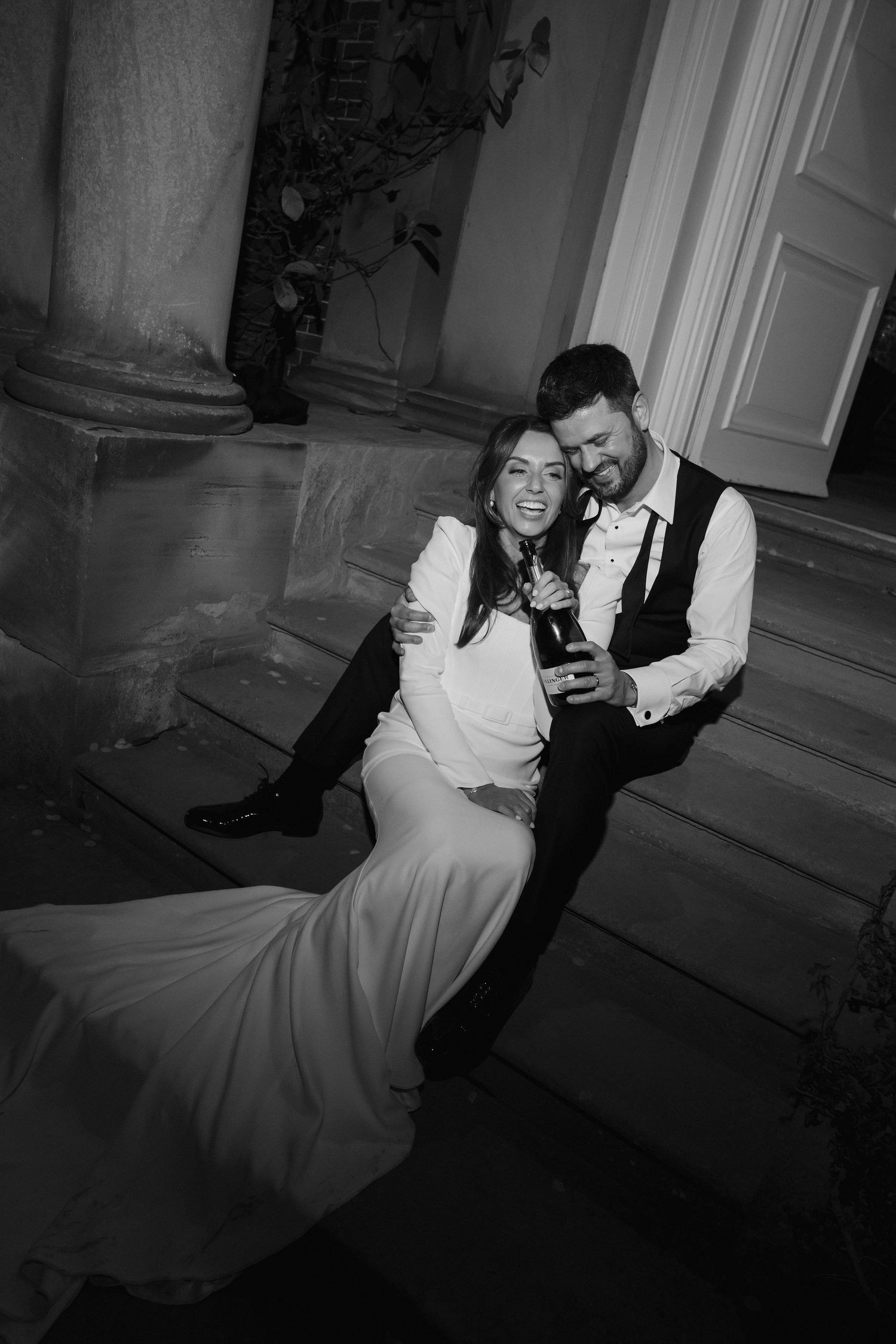 bride and groom sat on steps laughing and holding a bottle of champagne outside Iscoyd park at night