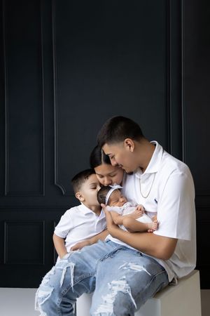 Intimate family moments captured against dark paneled wall in white outfits.