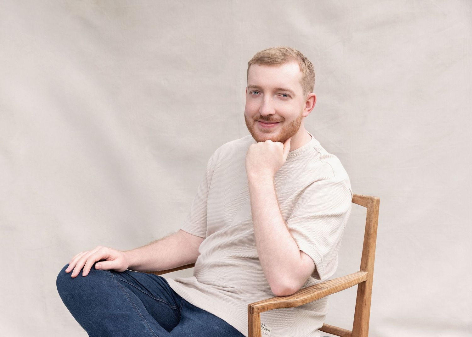 Person in white shirt sits casually in wooden chair against neutral background in studio setting.