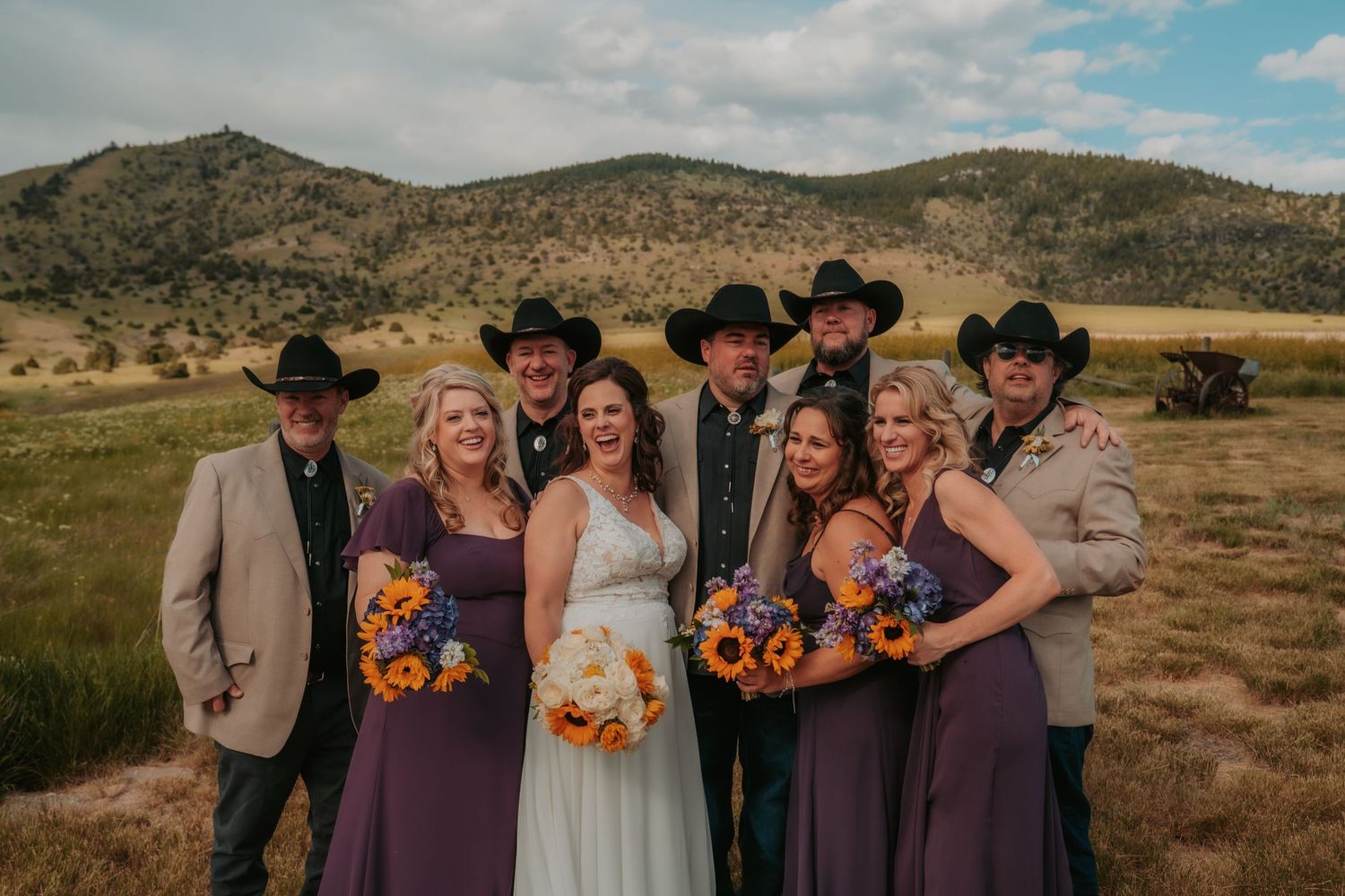 Wedding party in Western attire poses in front of scenic mountain landscape with sunflowers and purple dresses.