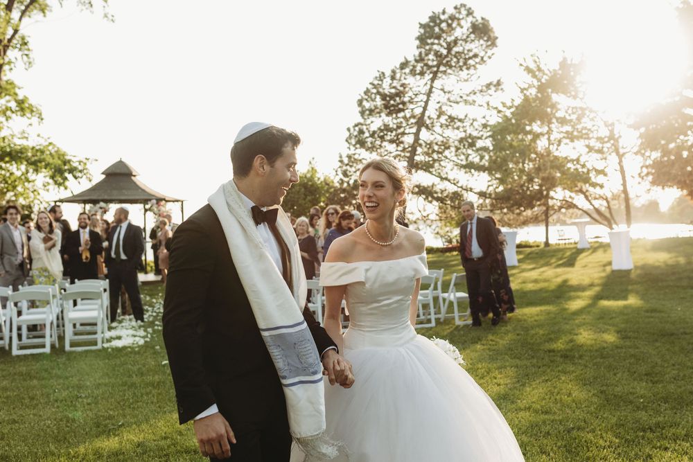 A wedding ceremony takes place on a sunlit lawn with white chairs and guests gathering near a gazebo.