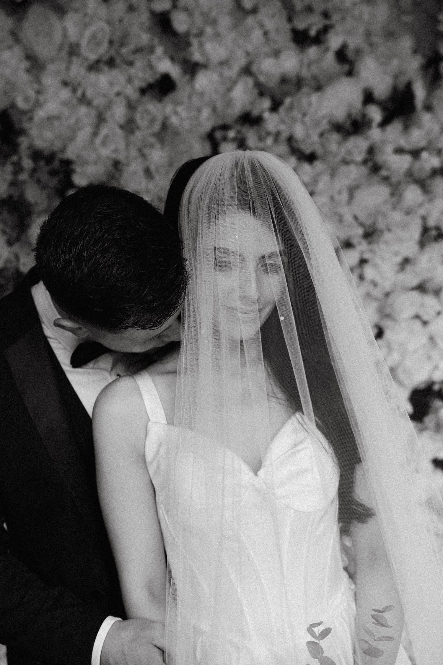 Black and white portrait of a bride in a sweetheart neckline dress with flowing veil against floral backdrop.