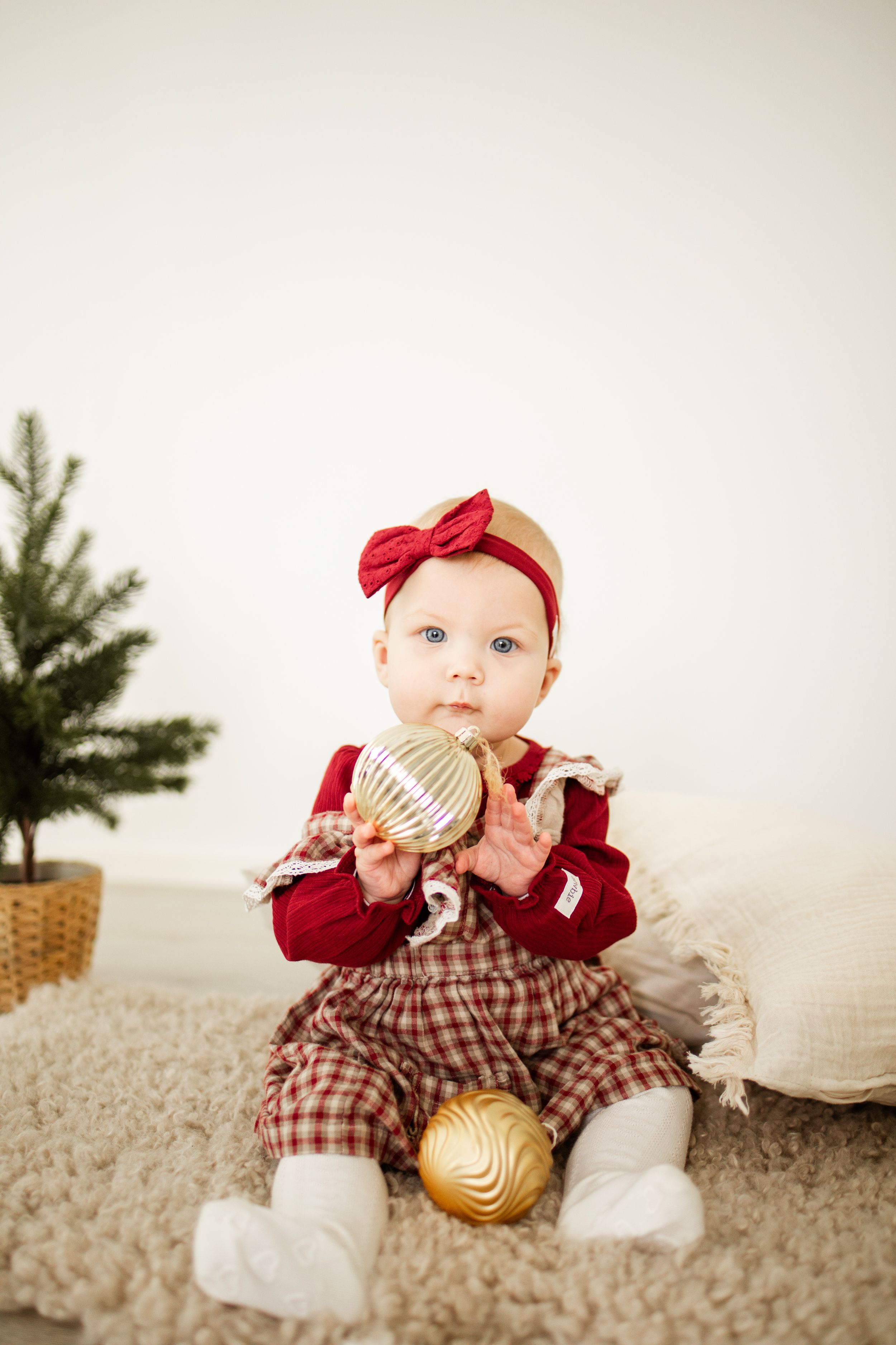 Baby in red and plaid holiday outfit sits on plush carpet with gold ornaments near small Christmas tree in background.