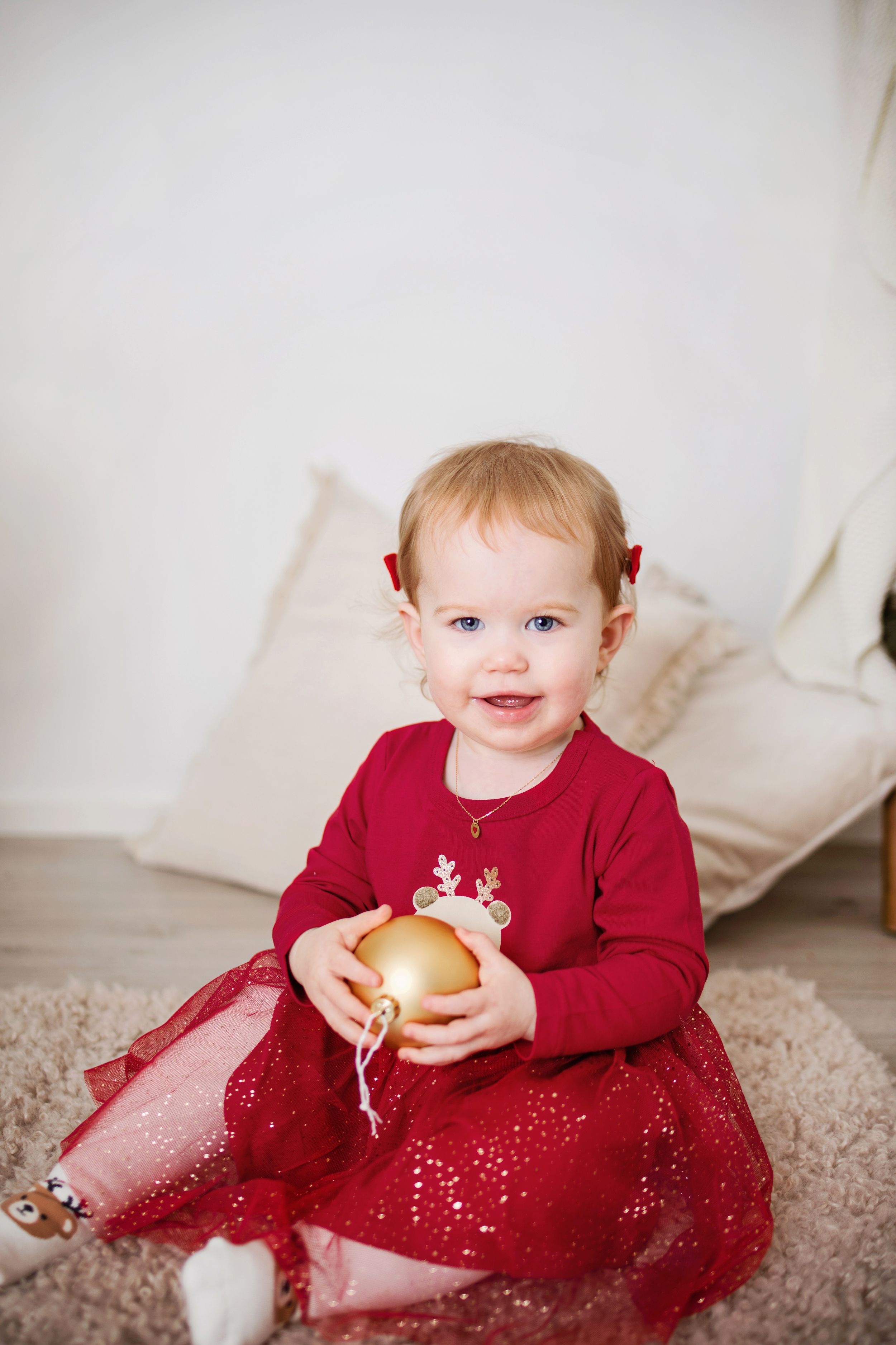 Toddler in a red dress sits on cream carpet playing with a golden ornament in four sequential photos.