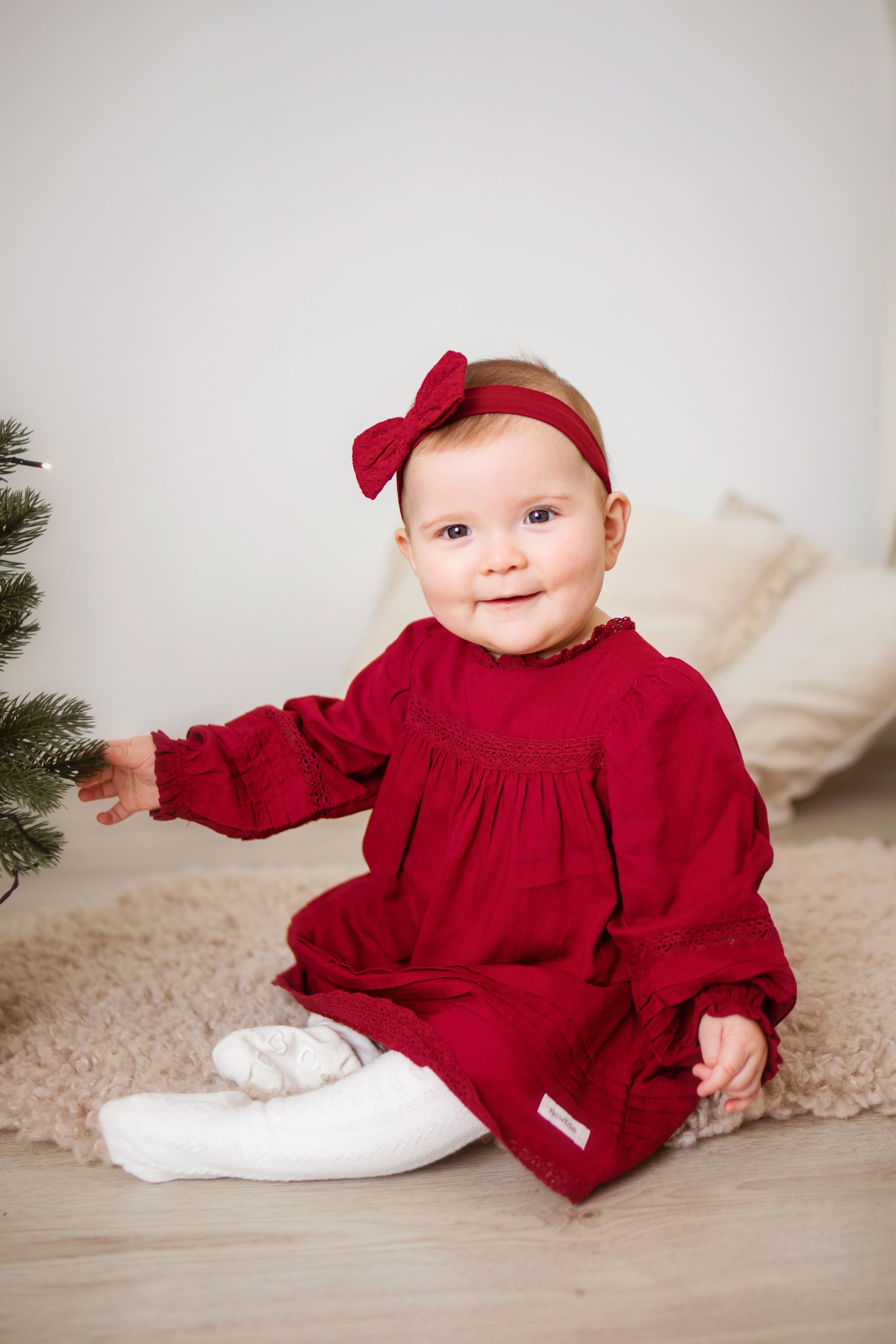 Baby in red velvet holiday dress and white socks sitting on cream carpet with evergreen branches in frame.