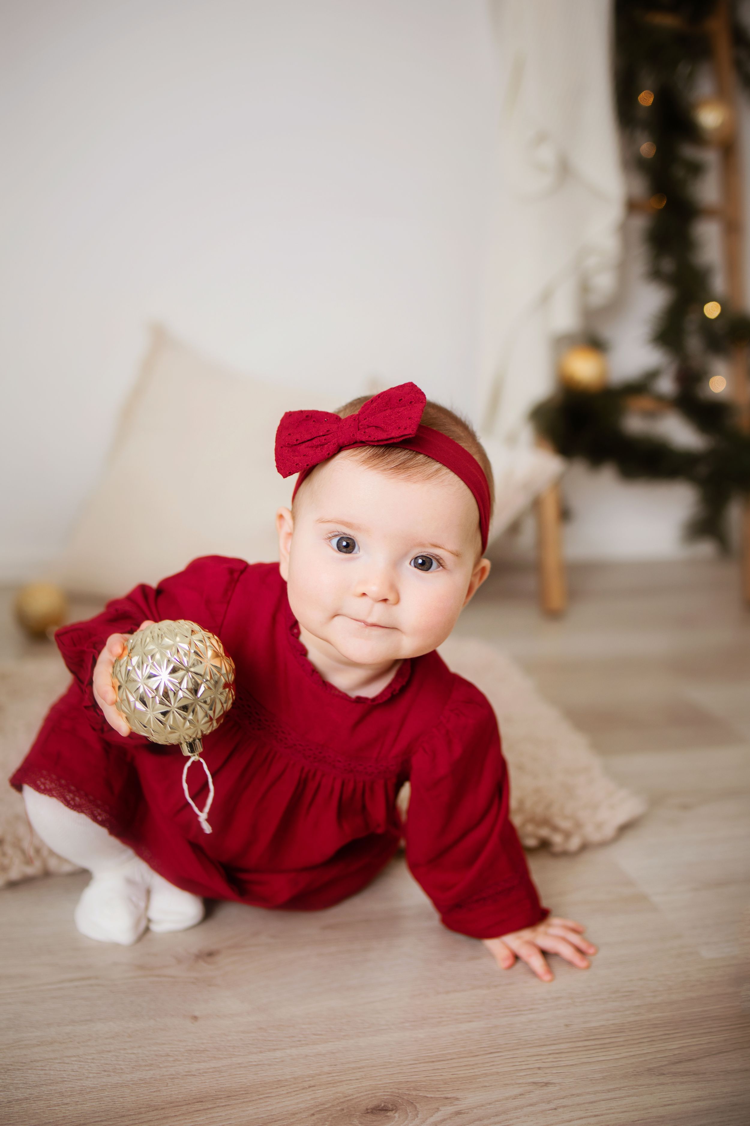 Baby in red velvet outfit crawls on hardwood floor with gold holiday decorations in background.