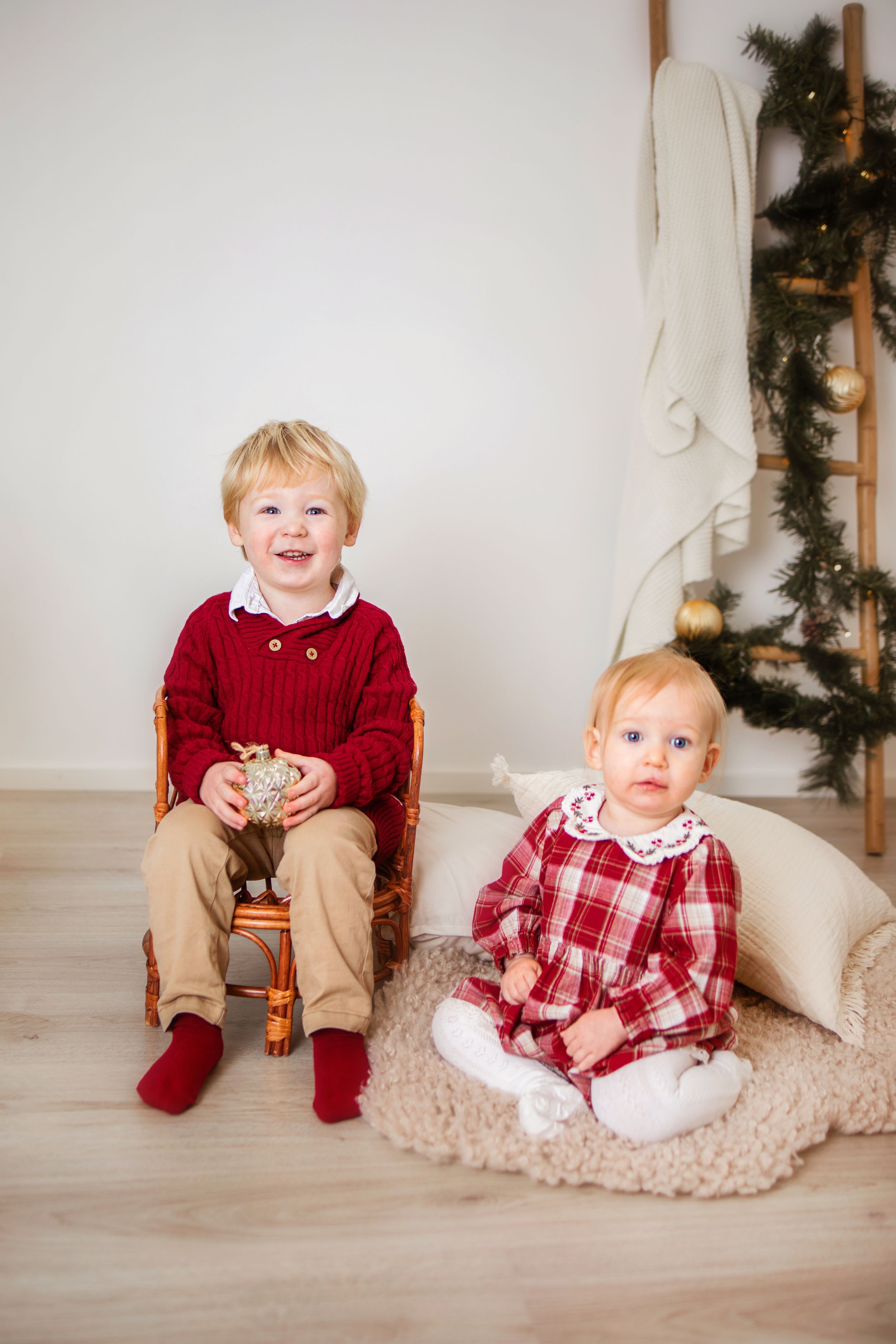 Child in festive holiday attire sits on cream-colored rug with Christmas decorations in red and plaid clothing.