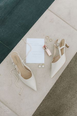 Elegant wedding flat lay featuring white slingback heels, pearl necklace, invitation card, and delicate jewelry on stone surface.