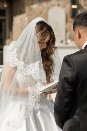 A bride in a lace-trimmed veil is seen from behind in soft lighting during an elegant wedding ceremony.
