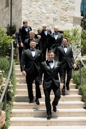 Groomsmen in classic black tuxedos and bow ties descend stone stairs at an elegant outdoor venue.