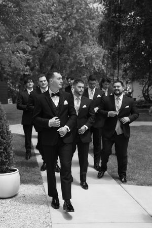 Groomsmen in matching black tuxedos and bowties walk together in a line while smiling and laughing outdoors.
